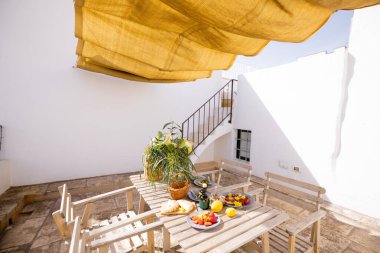 Beyaz bir terasta Rustik ahşap masa. Taze Akdeniz yemekleri, limonlar, çiçekler ve zeytinyağı ile altın sarısı bir güneş gölgesinin altında. Sıcak ve rahat yaz havası