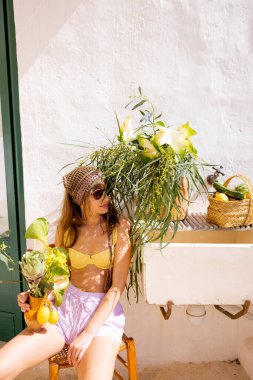 Yazlık kıyafetli bir kadın elinde bir buket limon ve enginar tutuyordu. Hasır sepet ve çiçeklerle kırsal bir lavabonun yanında çıplak ayakla oturuyordu. Akdeniz yaşam tarzı ve doğal güneş ışığı