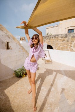 Kaygısız çıplak ayaklı kadın, güney İtalya 'da sıcak bir çatıda güneşlik tutarken şakacı bir şekilde gülümsüyor. Kırsal cazibe, sabah ışığı ve yaz havası.