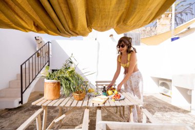 Kırsal bir çatıda altın bir güneşliğin altında yaz brunch 'ı düzenleyen bir kadın. Taze meyve, zeytinyağı, yerel ekmek ve arka planda neşeli beyaz bir köpek olan rahat bir atmosfer.