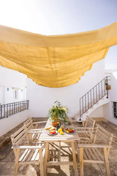 Beyaz bir terasta Rustik ahşap masa. Taze Akdeniz yemekleri, limonlar, çiçekler ve zeytinyağı ile altın sarısı bir güneş gölgesinin altında. Sıcak ve rahat yaz havası