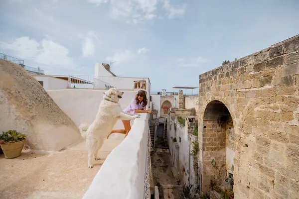 Bir kadın ve onun büyük beyaz köpeği çatıdaki çıkıntıdan eğilip aşağıdaki tarihi İtalyan caddesine bakıyorlar. Güneşli bir köyde huzurlu bir bağ anı