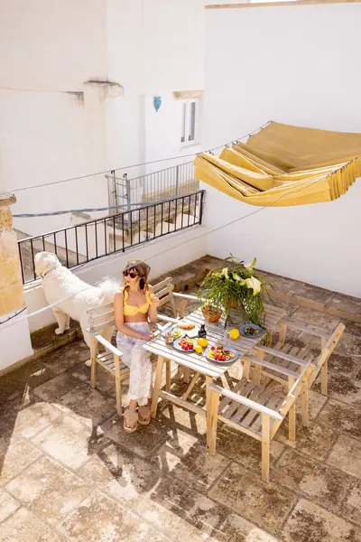 Güney İtalya 'da kırsal çatı terasında taze meyve ve kahveyle yavaş yavaş kahvaltı yapan bir kadın. Yaz ışığı, kaktüs bitkileri ve tüylü köpek dingin sabah havasını tamamlıyor.