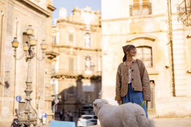 Bir kadın ve beyaz köpeği tarihi bir Sicilya meydanında huzurlu bir sabah yürüyüşü yapıyorlar. Görüntü, yavaş seyahat, arkadaşlık ve kültürel mirasın takdir edildiği hissini aktarıyor.