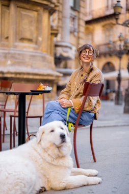 Gülümseyen bir kadın köpeğiyle birlikte açık bir kafede gevşeyip Sicilya dondurmasının tadını çıkarırken telefonunu kontrol ediyor. Fotoğraf dijital göçebe yaşam tarzını, yavaş yaşamanın neşesini ve evcil hayvanı gösteriyor.