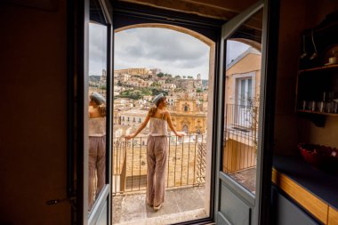 Panoramik manzaralı bir balkonda, barok çatıları ve Sicilya 'nın tepedeki tarihi kilisesi üzerinde duran bir kadın. Cam kapıdaki yansıma, yumuşak ışık ve sakin atmosfer