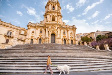 Kadın, büyük beyaz köpeğini Modica, Sicilya 'daki San Giorgio Katedrali' nin büyük merdivenlerinden geçerken parlak mavi bir gökyüzünün altında yürütür.