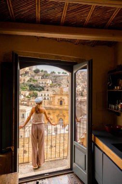 Panoramik manzaralı bir balkonda, barok çatıları ve Sicilya 'nın tepedeki tarihi kilisesi üzerinde duran bir kadın. Cam kapıdaki yansıma, yumuşak ışık ve sakin atmosfer