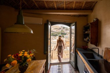 Sicilya 'nın Modica kentinin büyüleyici çatılarına ve tarihi Barok mimarisine bakan genç bir kadın balkonda duruyor. Açık pencereden yansıyan o huzurlu yamacı sessizce alır.