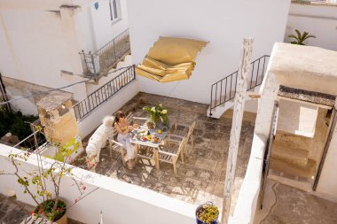 Güney İtalya 'da kırsal çatı terasında taze meyve ve kahveyle yavaş yavaş kahvaltı yapan bir kadın. Yaz ışığı, kaktüs bitkileri ve tüylü köpek dingin sabah havasını tamamlıyor.