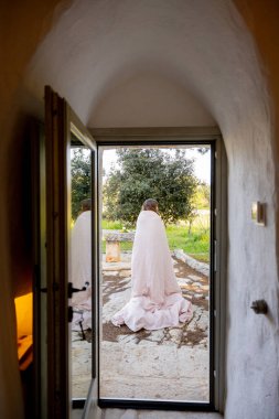 Çıplak ayaklı bir kadın, güneşin doğuşunda beyaz bir trullo evinin önünde pembe bir yorgana sarılmış. Sıcaklık, dinginlik ve kırsal bir günün huzurlu başlangıcı.