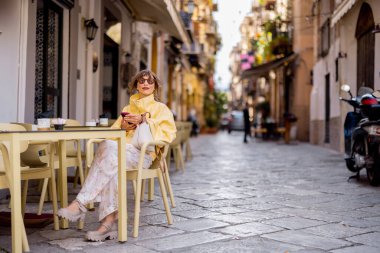 Sarı ceketli bir kadın, Palermos tarihi merkezinde bir kafede otururken telefonunu kontrol ediyor. Bu fotoğraf dijital göçebe yaşam tarzını, şehir estetiğini aktarır.
