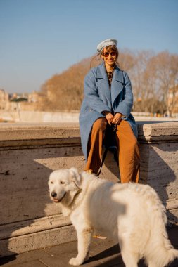 Roma 'da güneşli bir kış yürüyüşü sırasında neşeli bir an. Tiber 'in yanındaki taş bir duvarda, mavi bir paltoyla sarmalanmış, tüylü beyaz köpeğimle altın ışığın tadını çıkarıyorum.