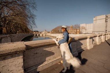 Mavi ceketli bir kadın, büyük beyaz köpeğini bir Roma köprüsünde kucaklıyor. Açık bir kış gökyüzünün altında St. Peters Bazilikası 'na bakıyor.