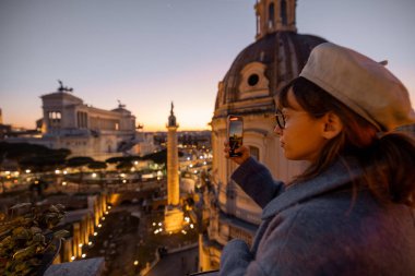 Mavi ceketli kadın günbatımında Roma siluetini yakalıyor. Arka planda Trajans Sütunu ve Vittoriano var. Manzaralı bir çatıda sıcak bir akşam ışığında parlıyor.
