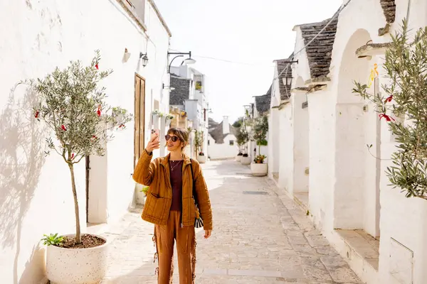 Gülümseyen kadın Alberobello, İtalya 'da geleneksel beyaz trulli evlerle dolu güneşli bir sokakta selfie çekiyor. Onu çevreleyen saksı zeytin ağaçları ve tarihi cazibe
