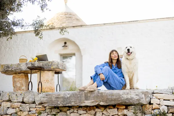 Mavi elbiseli bir kadın, geleneksel beyaz bir evin önünde, büyük beyaz köpeğinin yanında taş basamaklarda oturuyor. Görüntü, kırsal yaşamın sıcaklığını, güvenini ve dinginliğini aktarır.