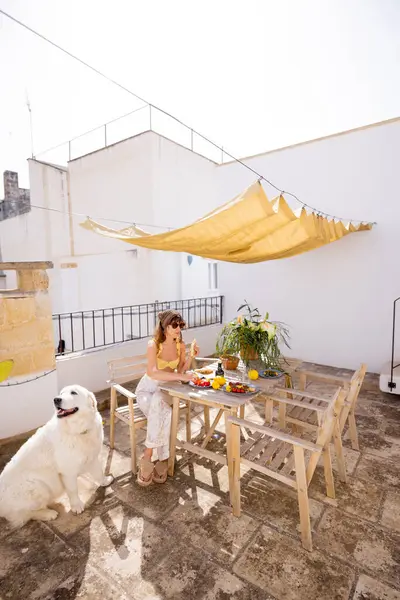 Güney İtalya 'da kırsal çatı terasında taze meyve ve kahveyle yavaş yavaş kahvaltı yapan bir kadın. Yaz ışığı, kaktüs bitkileri ve tüylü köpek dingin sabah havasını tamamlıyor.