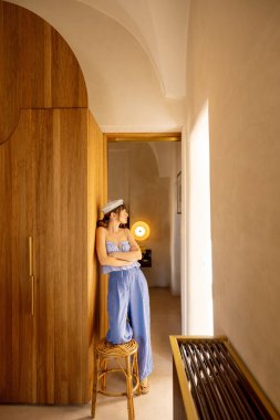 Açık mavi elbiseli ve bereli bir kadın yüksek tavanlı, güneşli bir koridorda ahşap bir duvara yaslanıyor. Huzurlu güney İtalya iç mimarisi
