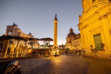 Roma 'nın ikonik Trajans Sütunu ve Altare della Patria' nın akşam manzarası, alacakaranlık gökyüzüne karşı aydınlandı, harabeler, kiliseler ve kaldırım taşı sokaklarla çevrili.