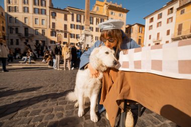 Mavi paltolu bir kadın, Roma meydanındaki bir kafede beyaz köpeğini kucaklıyor. Roma 'nın kalbinde huzurlu bir sevgi anı yakalıyor.
