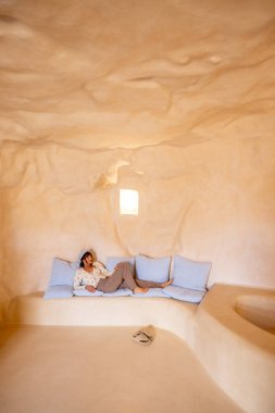 Sicilya 'da, Modica' da oyulmuş bir mağara evinde çıplak ayakla mavi yastıklarda dinlenen bir kadın. Topraksı tonlar ve minimalizm geleneksel Akdeniz 'de barışçıl ve topraklayıcı bir atmosfer yaratıyor.