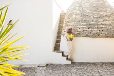 Çiçek kıyafetli ve pembe bereli bir kadın geleneksel konik trullo çatısı olan beyaz taş bir ev boyunca yürür, elinde yumuşak güneş ışığı altında bir buket parlak sarı ranunculus taşır.