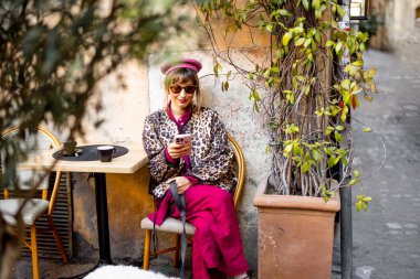 Leopar desenli ceketli ve pembe elbiseli şık bir kadın Roma kafesinin önünde telefonuna bakıyor. Bu görüntü modern şehir yaşam tarzını ve sokak modasını yansıtıyor.