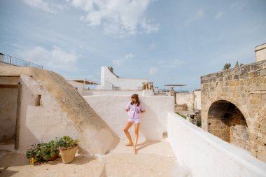 Genç bir kadın ve tüylü beyaz köpeği tarihi taş duvarlar ve saksı bitkileriyle çevrili bir Akdeniz çatısı üzerinde duruyorlar. Güney İtalya 'da sakin ve güneşli bir an