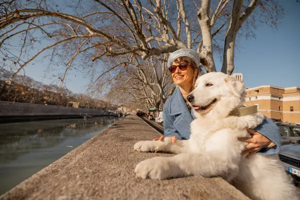 Roma 'daki Tiber Nehri' nin yanında bir kadın ve tüylü beyaz köpeği çıplak çınar dalları altında manzarayı ve kış güneşini paylaşıyor. Ebedi şehrin sakin güzelliğini kucaklıyor.