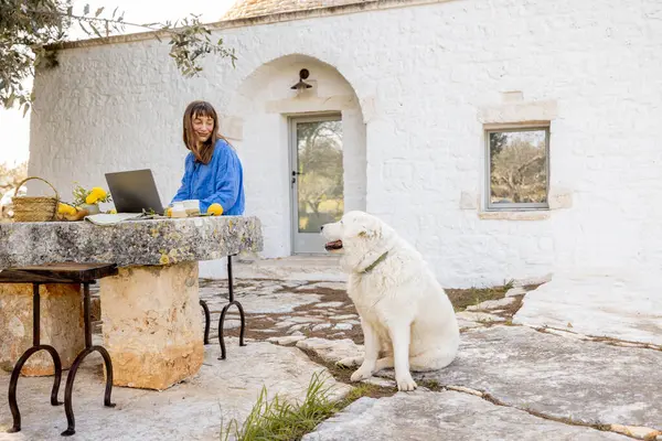 Bir kadın köpeğiyle birlikte trullo evinin önündeki taştan bir masada dizüstü bilgisayarıyla çalışıyor. Konsept uzak çalışmaları, dijital göçebe yaşam tarzını ve barışçıl doğa bağlantısını yakalıyor