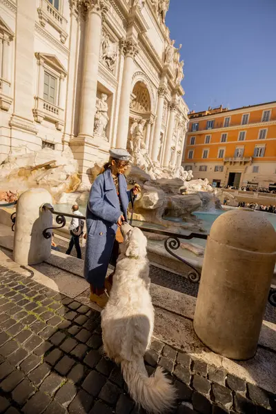 Bir kadın ve tüylü beyaz köpeği Roma 'daki Trevi Çeşmesi' nin orada güneşli bir anın tadını çıkarıyorlar. Huzurlu, iç açıcı bir yoldaşlık ve seyahat sahnesi.