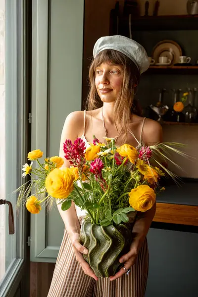 Yumuşak bereli genç bir kadın pencerenin kenarında duruyor. Elinde, ılık doğal ışıkla yıkanmış canlı sarı ve mor bir çiçek buketi var. Huzurlu, zamansız bir sadelik ve cazibe sahnesi.