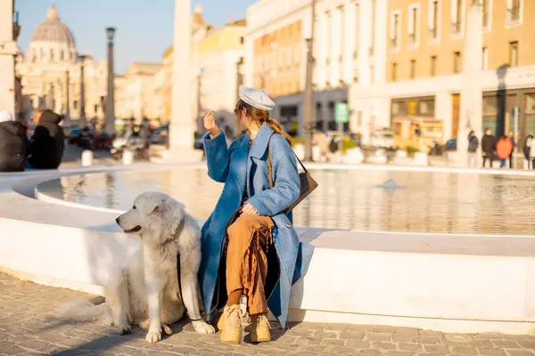 Mavi ceketli gülümseyen bir kadın St. Peter Bazilikası yakınlarındaki büyük beyaz köpeği yanında oturan modern bir çeşmeye bozuk para atıyor. Roma 'da umudun sembolü ve neşeli seyahat anları