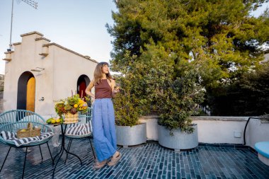 Gülümseyen kadın kiremitli terasta çıplak ayakla yürüyor enginarlar ve limonlarla dolu büyük bir buket tutuyor yeşil ve kırsal cazibeyle çevrili huzurlu güneşli bir günün tadını çıkarıyor.