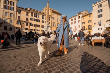 Mavi ceketli şık bir kadın, pofuduk beyaz köpeğini, yerli ve turistlerle dolu güneşli bir Roma meydanında gezdiriyor.