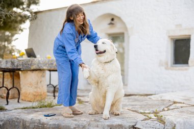 Mavi elbiseli gülümseyen bir kadın geleneksel beyaz trullo evinin dışında tüylü beyaz köpeğiyle patisini titretmekte, kırsal bir ortamda sıcaklık ve arkadaşlık saçmaktadır.