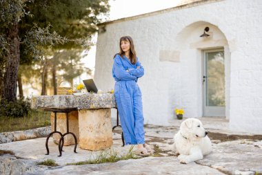 Mavi tulumlu bir kadın, köpeği ve laptopuyla geleneksel bir trullo 'nun önünde güvenle duruyor. Konsept iş ve kırsal yaşam arasındaki dengeyi gösteriyor.