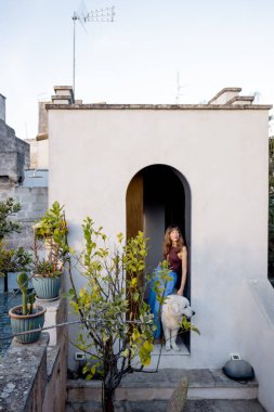 Bir kadın ve beyaz köpeği saksı bitkileri ve taş mimarisi ile çevrili bir çatı bahçesine çıkarak huzurlu Akdeniz yaşam tarzını ve doğayla bağlantısını anımsatıyor.