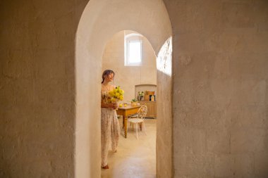 Bir kadın taştan bir mutfaktaki ahşap bir masada sarı çiçekler düzenliyor. Sahne basitlik, gelenek ve konforlu Akdeniz yaşamını iç mekan veya yaşam tarzı kullanımı için mükemmel bir şekilde yansıtıyor