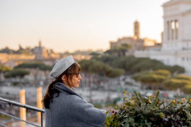 Mavi paltolu ve bereli bir kadın Roma Forumu ve Altare della Patria üzerinde gün batımını seyrediyor. Tarihin ve seyahatin ebedi güzelliği