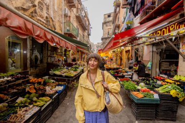 Sarı ceketli bir kadın Sicilya 'da canlı bir yerel markette geziniyor, taze ürünlerle çevrili. Görüntü otantik seyahat, kültürel dalma ve yavaş yaşam tarzını aktarıyor. 