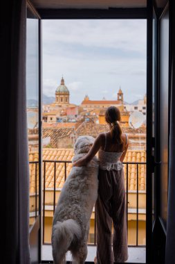 Bir kadın ve tüylü beyaz köpeği, huzurlu bulutlu bir sabahta Sicilya, Palermo 'nun çatılarına ve kubbelerine bakan balkonda sakin bir anı paylaşıyor.