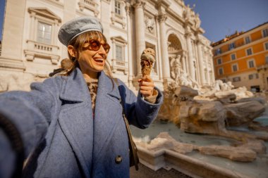 Mavi ceketli gülen kadın, Roma Trevi Çeşmesi 'nin önünde dondurmasının tadını çıkarıyor. Ebedi Şehrin kalbinde tatlı ve güneşli bir anı kutluyor.