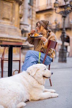 Gülümseyen bir kadın köpeğiyle birlikte açık bir kafede gevşeyip Sicilya dondurmasının tadını çıkarırken telefonunu kontrol ediyor. Fotoğraf dijital göçebe yaşam tarzını, yavaş yaşamanın neşesini ve evcil hayvanı gösteriyor.