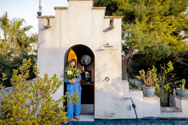 Elinde canlı bir buket tutan genç bir kadın yemyeşil bitkiler, kır taşı ve Akdeniz cazibesiyle çevrili güneşli bir terasa adım atıyor.