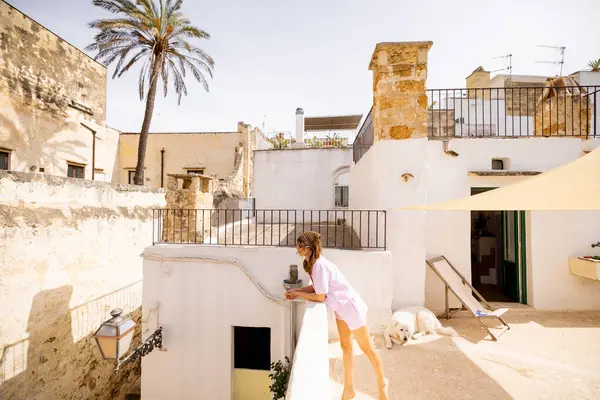 Akdeniz taş duvarları, palmiye ağaçları ve güney İtalya 'da dinlenen beyaz köpekli güneşli bir çatı manzarasının keyfini çıkaran bir kadın.
