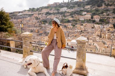 Büyük beyaz köpekli kadın, Sicilya, Modica üzerindeki panoramik manzaranın tadını çıkarıyor. Terracotta çatıları ve barok mimari ile çevrili mükemmel bir bağlantı, huzur ve yavaş seyahat anı.