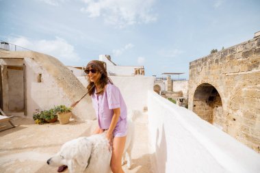 Genç bir kadın ve tüylü beyaz köpeği tarihi taş duvarlar ve saksı bitkileriyle çevrili bir Akdeniz çatısı üzerinde duruyorlar. Güney İtalya 'da sakin ve güneşli bir an