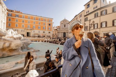 Şık bir kadın Roma ikonik Trevi Çeşmesi 'nin önünde dondurmayı seviyor, hareketli meydanın güneş ışığını ve hareketli atmosferini emiyor.